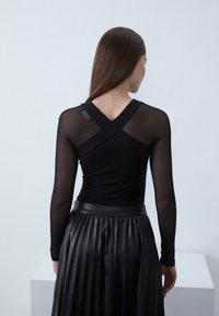 Juoda ilgomis rankovėmis palaidinė su permatomomis tinklelio rankovėmis, su kryžminio dizaino nugaroje. Derinama su juoda plisuota netikros odos suknele.