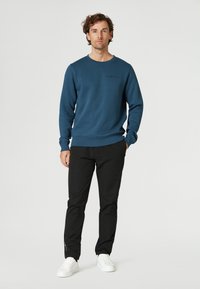 Sudadera de forro polar azul con mangas largas y puños acanalados, combinada con pantalones negros y zapatillas blancas, de pie frente a un fondo liso.