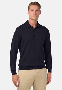 Jūras spēku tumši zils garā piedurkne polo krekls no teksturēta auduma, ar klasisku apkakli un trīs pogu aizdari, apvienots ar gaišas krāsas biksēm.