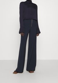 Pantaloni larghi blu marino con dettaglio zip frontale. Abbinati a un top a maniche lunghe con collo alto a coste, caratterizzato da maniche a campana.