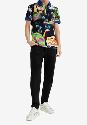 Persoon draagt zwarte spijkerbroek, witte sneakers met zwarte krabbels en een kleurrijk poloshirt met grafische kunst en tekstelementen.