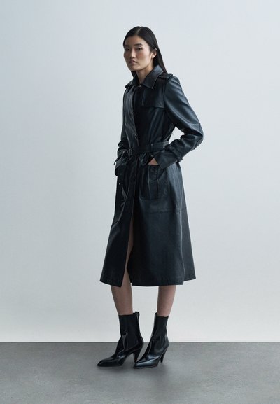 Czarny skórzany płaszcz typu trencz z paskiem w talii, zapięciem na guziki i dwoma przednimi kieszeniami. Dopasowany fason o długości do połowy uda, zestawiony z czarnymi botkami za kostkę.