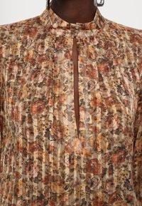 Blouse à motif floral avec des tons chauds marron, orange et beige, col montant fermé par trois boutons argentés et ouverture en forme de trou de serrure à l'avant.