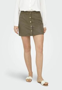 Mini falda de ante verde oliva con corte recto, con cierre frontal de botones y textura suave. Combinada con sandalias planas blancas.