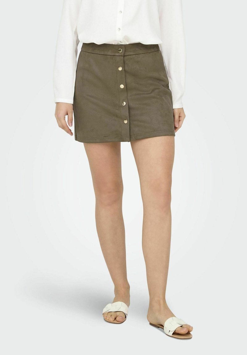 Mini falda de ante verde oliva con corte recto, con cierre frontal de botones y textura suave. Combinada con sandalias planas blancas.