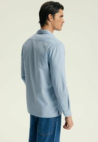 Jasnoniebieska koszula z długim rękawem, o prostym kołnierzyku, z mankietami na guziki i bocznymi rozcięciami. Miękka tekstura, minimalistyczny design i dopasowany krój.