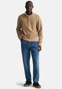Beige, langemouw pullover met een half-rits kraag, gecombineerd met losse blauwe jeans en zwarte sneakers met bruine zolen. Glad stoftextuur.