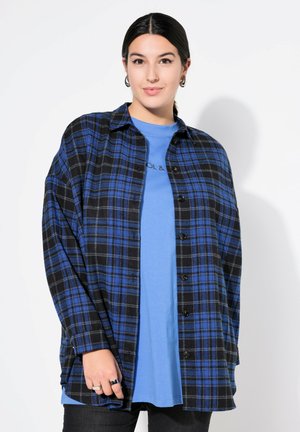 Zwarte en blauwe geruite blouse, knoopsgat, met kraag en een losse pasvorm, gedragen over een blauwe t-shirt. Model toont zwarte skinny jeans en ringen.