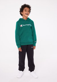 Felpa verde con cappuccio e tasca frontale, logo bianco in scritta, abbinata a pantaloni sportivi neri con un piccolo logo e sneakers bianche.
