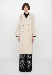 Beige double-breasted trench coat with large collar, gold buttons, and wide sleeves. Paired with black pants featuring white floral patterns.
