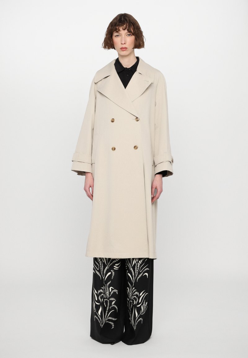 Beež kahe nööbiga trench coat suure kraega, kuldsete nööpidega ja laia varrukatega. Kombineeritud mustade pükstega, millel on valged lillemustrid.