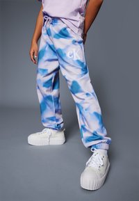 Persoon in blauwe en witte tie-dye sweatpants met "cK" logo, witte platform sneakers en een lichtpaarse shirt tegen een effen grijze achtergrond.
