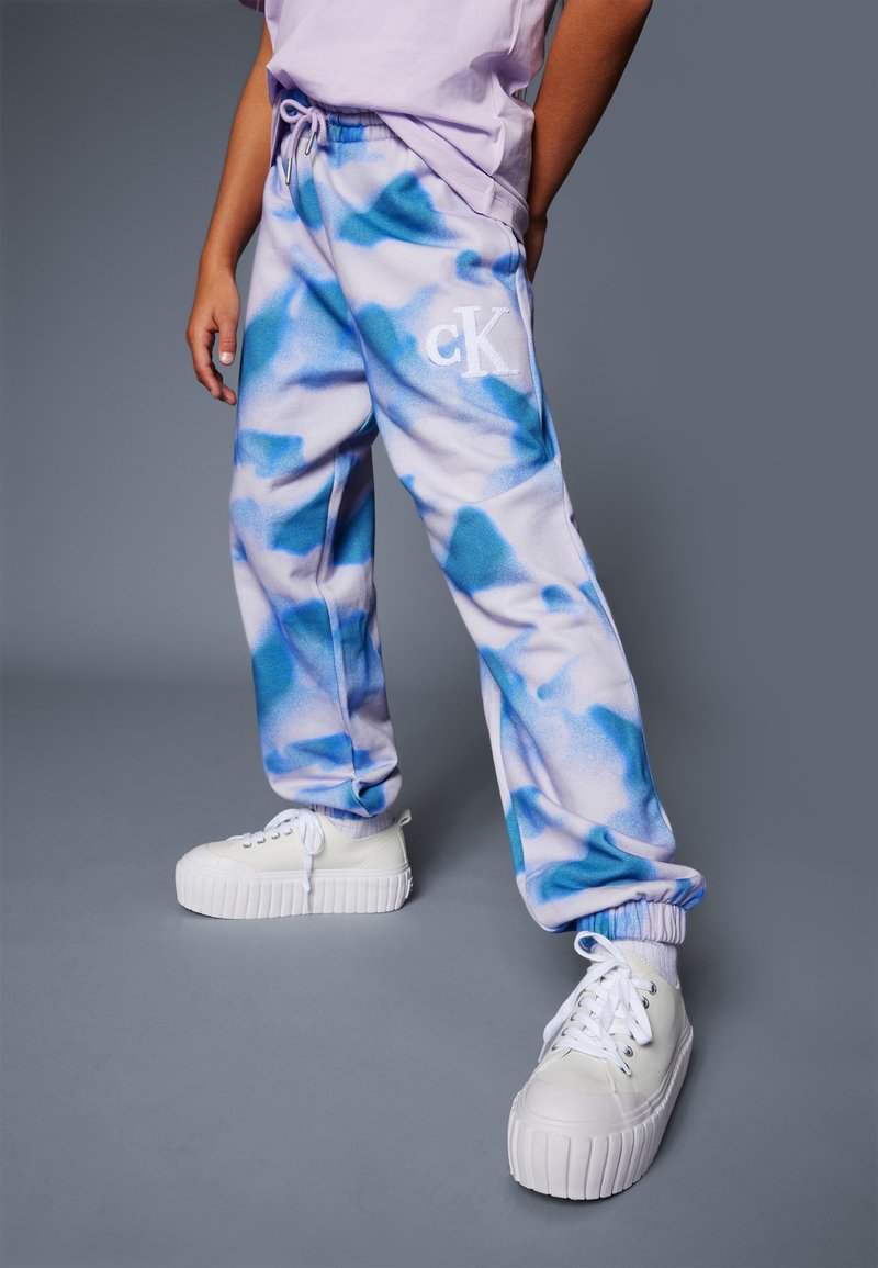 Persoon in blauwe en witte tie-dye sweatpants met "cK" logo, witte platform sneakers en een lichtpaarse shirt tegen een effen grijze achtergrond.