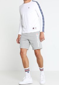 Biała koszula z długimi rękawami z granatowymi akcentami, szare szorty z białym napisem "Champion" oraz białe trampki z czerwono-niebieskimi paskami na skarpetkach.