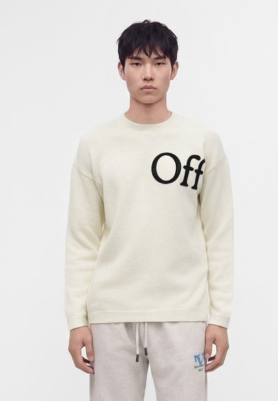 Jeune homme aux cheveux courts et noirs, portant un pull crème avec le texte "Off" et un pantalon de survêtement gris clair, debout devant un fond uni.