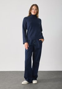 Pulover tricotat cu guler înalt de culoare bleumarin, cu mâneci lungi, asortat cu pantaloni din denim de culoare albastru închis, cu talie înaltă și croială lejeră. Tenisi albi completează acest look.