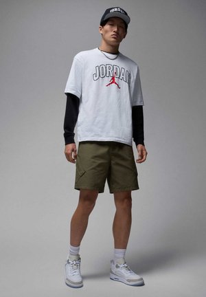 Biała koszulka z napisem "JORDAN" i czerwonym logo, czarna koszula z długim rękawem pod spodem, oliwkowe szorty oraz białe sneakersy z szarymi akcentami.
