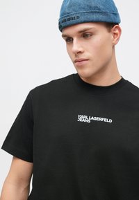 Jaunas vyras, vilkintis juodą marškinėlį su užrašu "Karl Lagerfeld Jeans" ir mėlyną džinsinį kepuraitę, žiūri šiek tiek žemyn ir šoną.