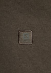 Brauner Stoff mit einer strukturierten, gewebten Oberfläche, versehen mit einem quadratischen Logo-Patch mit dem eingeprägten Schriftzug "GABBIANO", umrahmt von einem kontrastierenden Faden.