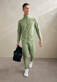 Maslinasto zelena trenirka s bijelim prugama, jakna na patentni zatvarač i elastični rukavi. Muškarac nosi crnu torbu za sportsku opremu i nosi bijele tenisice.