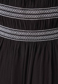 Svart plagg med texturerad tyg och samlade veck, accentuerad av horisontell vit broderi i geometriska mönster.