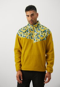 Gul fleece genser med en mønstret blå og gul øvre del. Har en halv glidelås foran og en teksturert, myk overflate.