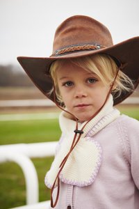 Brūna ādas kovboja cepure ar pītām lentēm, krēmkrāsas šalle ar violetām šuvēm, gaiši violeta džemperis, zaļas zāles fons.
