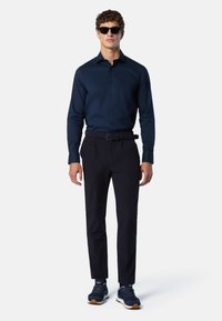 Camicia blu con bottoni, cintura scura, pantaloni blu slim-fit e sneakers blu con un motivo texture; modello che indossa occhiali da sole neri.