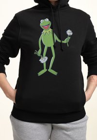 Haut à capuche noir avec un personnage de grenouille verte imprimé tenant des marguerites, avec un cordon de serrage au niveau de la capuche et des poignets et un ourlet côtelés.