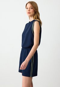 Completo sportivo senza maniche di colore blu navy con una vestibilità rilassata, caratterizzato da righe laterali bianche e verdi sui pantaloni. Texture morbida del tessuto.