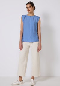 Blusa senza maniche azzurra chiara con scollatura rotonda, bottoni frontali e dettagli arricciati sottili, abbinata a pantaloni bianchi larghi e scarpe casual.