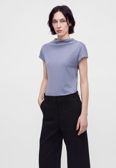 Femme aux cheveux courts et foncés porte un haut violet clair à manches courtes et un pantalon noir taille haute à jambes larges, debout devant un fond uni.