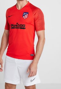 Hombre con una camiseta de fútbol roja del Atlético de Madrid y pantalones cortos blancos de Nike, de pie contra un fondo claro y liso.
