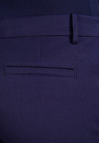 Närbild av mörkblå dressbyxor som visar en bakficka med bälte och detaljerad sömnad.