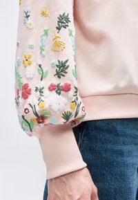 Μανίκι ροζ φούτερ με πολύχρωμα κεντητά λουλούδια, ριμπ μανσέτα και ένα χέρι που ακουμπά κοντά σε μπλε τζιν.