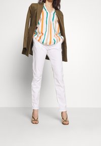 Femme portant un blazer olive posé sur les épaules, un chemisier à rayures à col en V, un pantalon blanc et des sandales à talons à brides olive, debout les mains dans les poches.