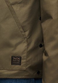 Χακί πράσινο μπουφάν με μαύρα κουμπιά, διαγώνια τσέπη και μαύρη τετράγωνη ετικέτα με χρυσό κείμενο "TAION" πάνω από μπλε τζιν από τζιν ύφασμα.