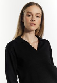 Black knit sweater with a V-neck and short collar. Features long sleeves and a smooth texture. Model shown from the chest up against a white background.