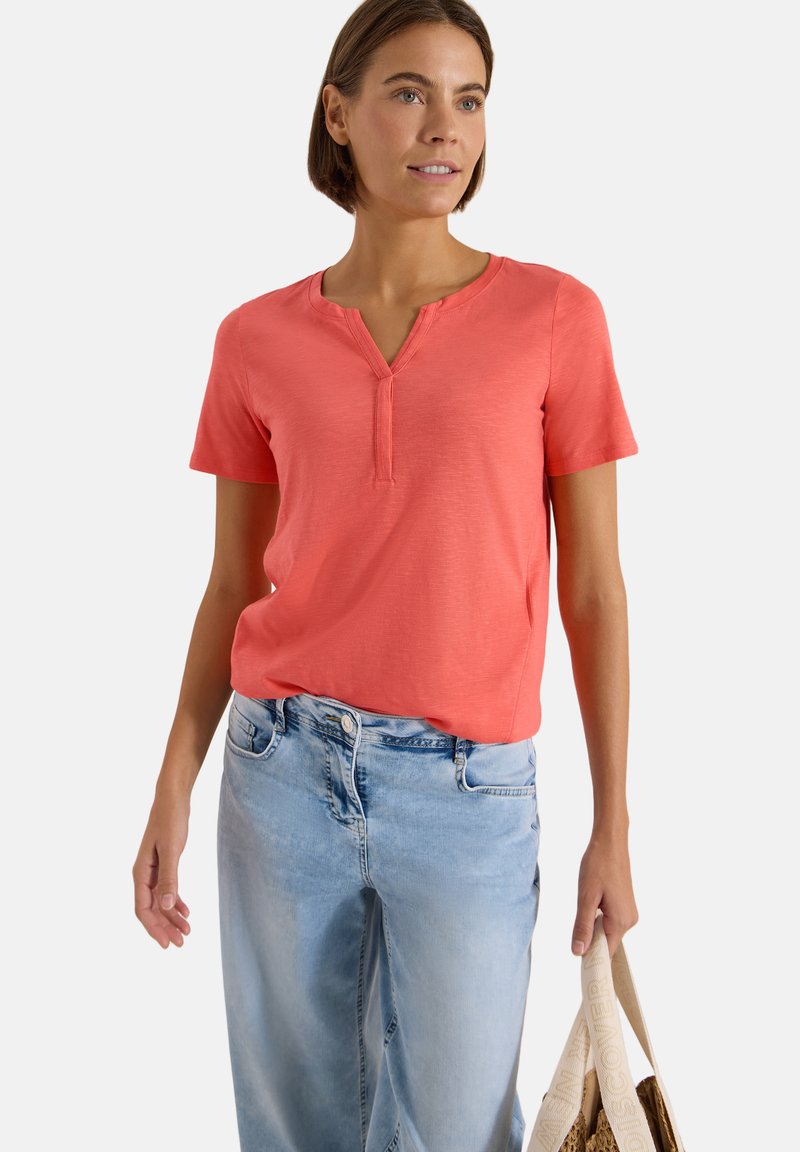 Femme portant un haut corail à manches courtes et un jean bleu clair, tenant un sac fourre-tout beige avec un texte imprimé sur la bandoulière.