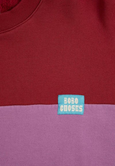 Gros plan sur un vêtement en tissu rouge et violet avec un patchwork de couleurs, comportant une petite étiquette bleue inscrite "BOBO CHOSES" cousue près de la couture entre les couleurs.