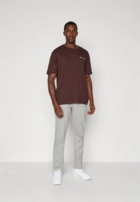 Rjava bombažna majica s kratkimi rokavi in majhnim logotipom, kombinirana s svetlo sivimi trenirkami in belimi supergami, stoječa na enobarvni podlagi.