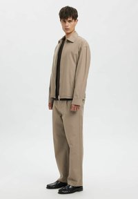 Beige lynlåsjakke over en mørk top, parret med løstsiddende beige bukser og sorte sko. Glat stof med minimal detaljering.