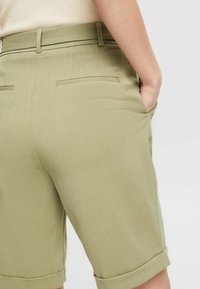 Shorts ajustés vert olive avec ourlets retroussés, poches latérales et deux poches arrière. Texture lisse, design ajusté, devant plat.