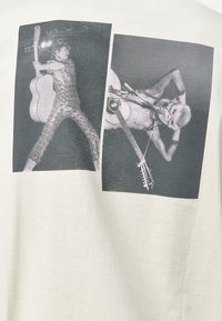 Lichte kleur T-shirt met daarop twee zwart-wit foto's van muzikanten die gitaar spelen, met verschillende poses en gedetailleerde achtergronden.