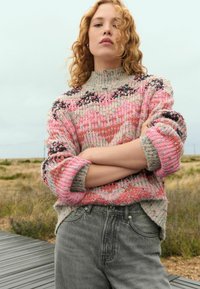 Pull en tricot multicolore avec un col haut, présentant des nuances de rose, de gris et des motifs. Assorti à un jean gris taille haute.