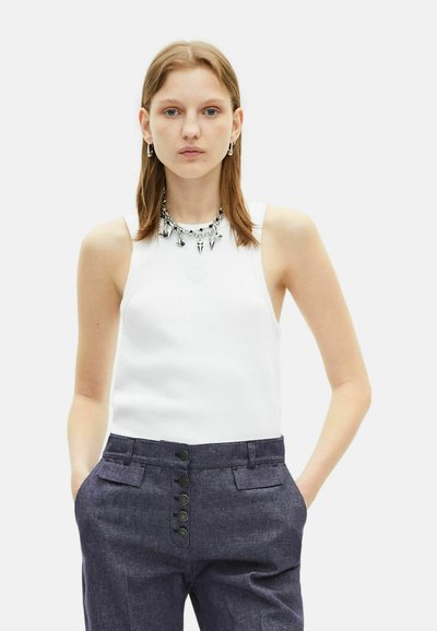 Witte mouwloze top met een geribbelde textuur, gecombineerd met donkerblauwe broek met knoopaccenten en zijzakken. Zilveren ketting met hangers.