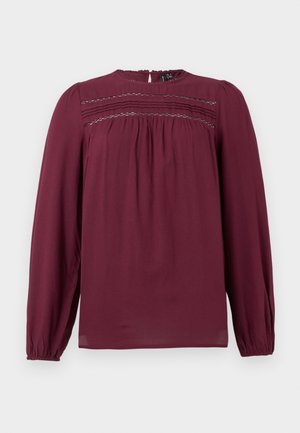 Burgundy blouse with long sleeves, round neckline, and gathered detailing at the chest. Smooth fabric with subtle texture and soft draping.