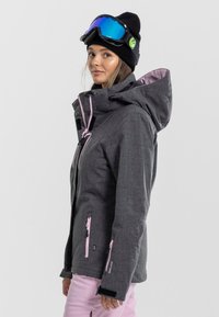 Grijze skijas met een capuchon, contrasterende roze ritsen en verstelbare manchetten. Gecombineerd met roze broek en een zwarte beanie en skibril.