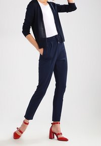 Navy blue relaxed-fit trousers with an elastic waistband, paired with a white T-shirt and a lightweight navy cardigan. Red high-heeled shoes.