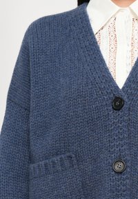 Sinine koorditud kardigan mustade nööbide ja tekstuurse kootud mustriga, kantud valge pitspluusiga, millel on terav krae.